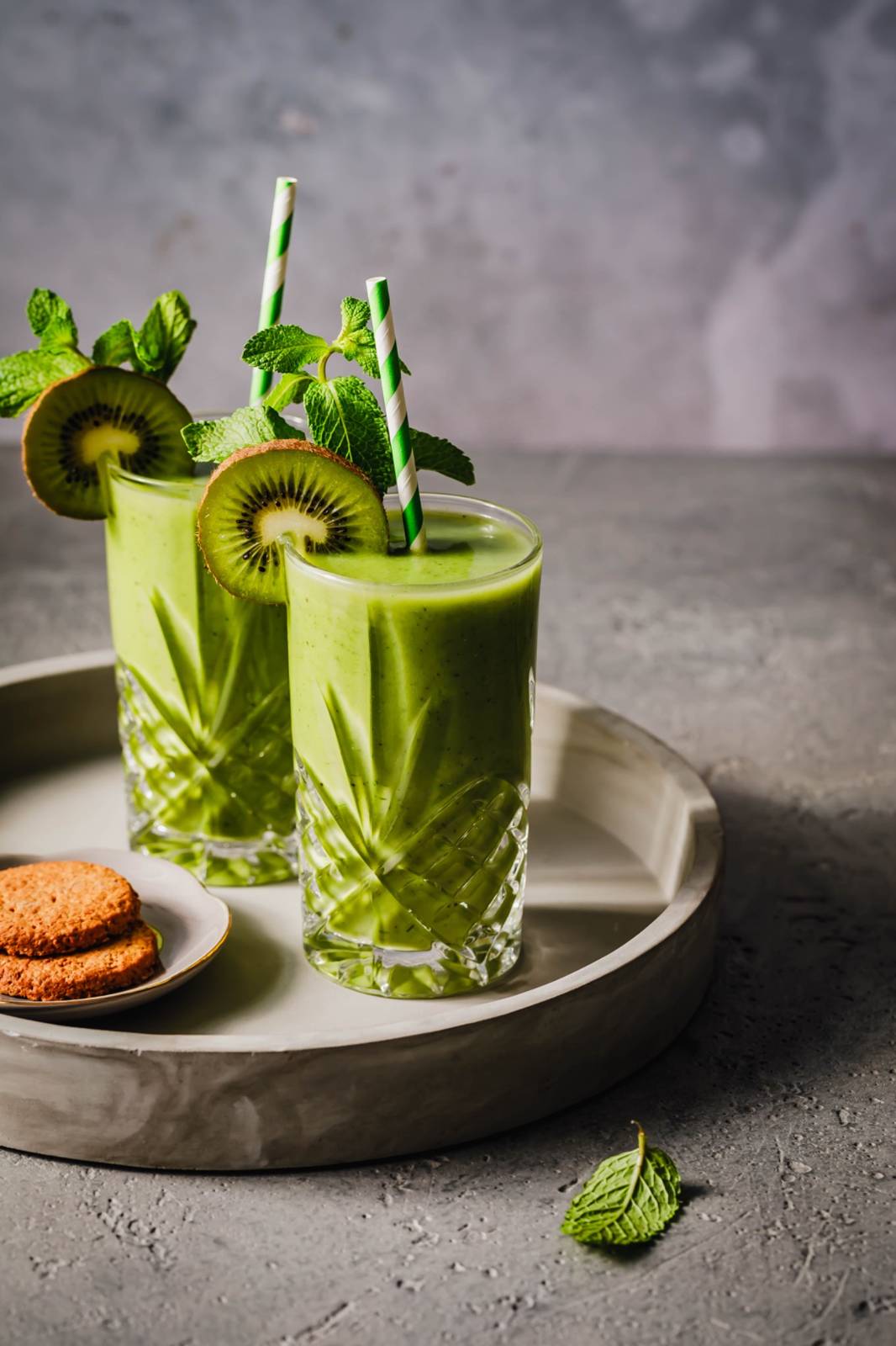 Zwei Gläser mit grünem Smoothie, garniert mit Minze, Kiwischeiben und gestreiften Strohhalmen auf einem Tablett, daneben ein kleiner Teller mit zwei Keksen vor einem grauen Hintergrund.
