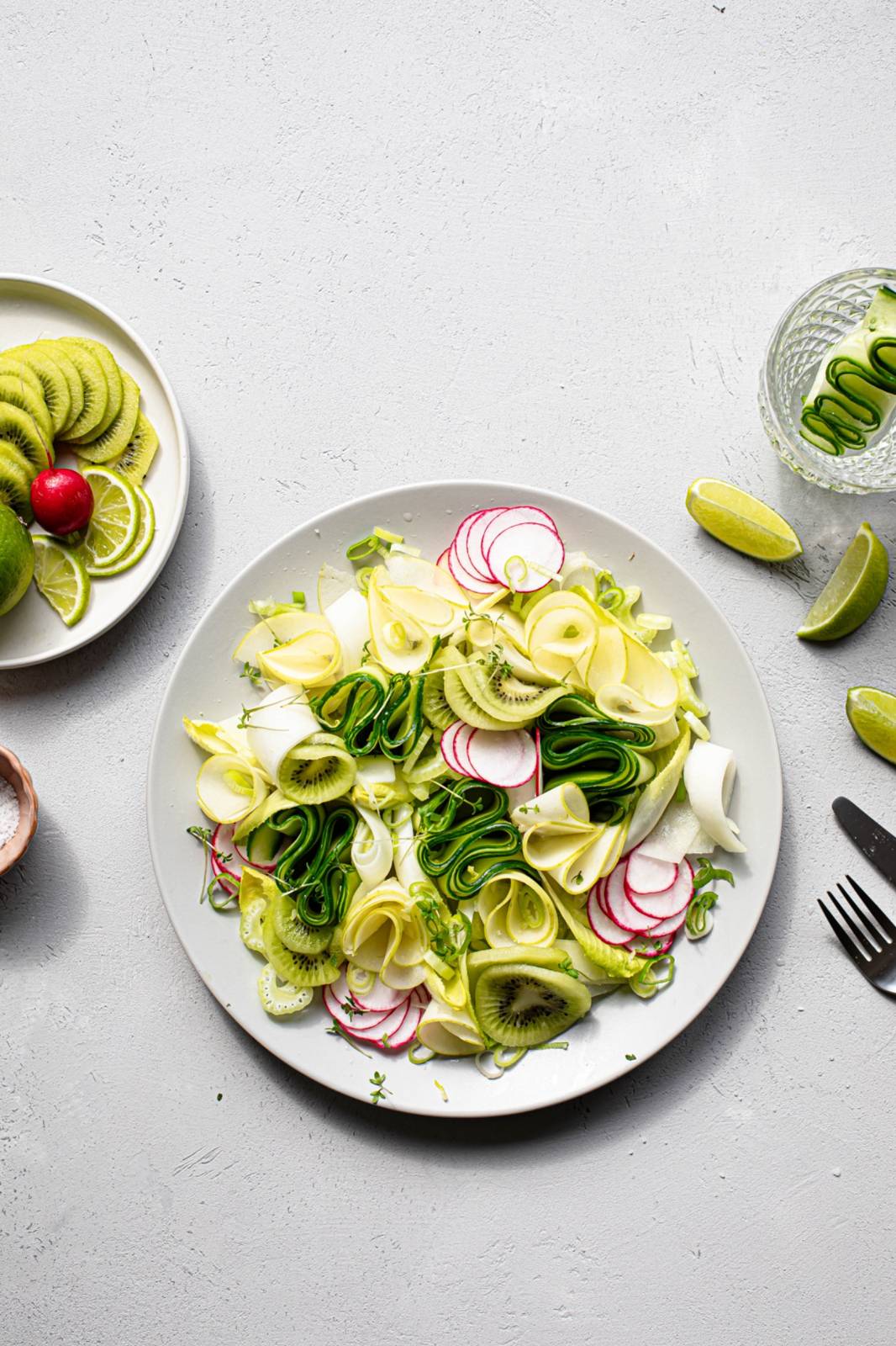 Ein Teller mit frischem Salat mit dünn geschnittenen Zucchini, Radieschen, Kiwi und Endivien, die in dekorativen Wirbeln angeordnet sind. Daneben ein Teller mit geschnittenen Limetten und Radieschen, Limettenspalten und Besteck auf einer hellen Unterlage.