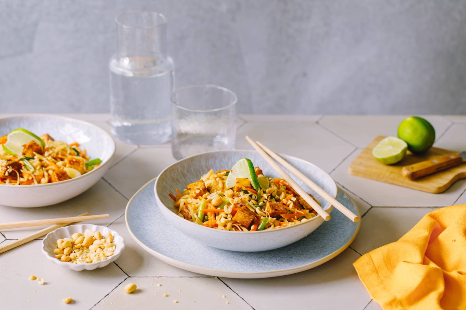 Eine Schüssel Pad Thai, garniert mit Limettenspalten, Erdnüssen und grünen Zwiebeln, steht auf einem Teller mit Stäbchen. Im Hintergrund stehen ein Glas Wasser, eine weitere Schüssel und ein Schneidebrett mit geschnittenen Limetten.