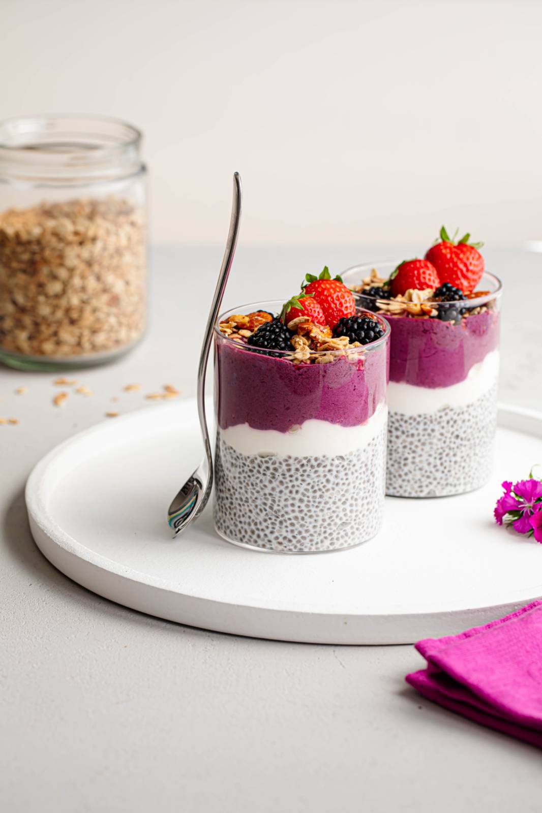 Zwei Gläser mit geschichtetem Chia-Pudding mit weißem Chia, lila Smoothie und Joghurt, garniert mit Erdbeeren, Brombeeren und Müsli. Ein Glas Granola und ein silberner Löffel stehen daneben auf einem weißen Tablett.