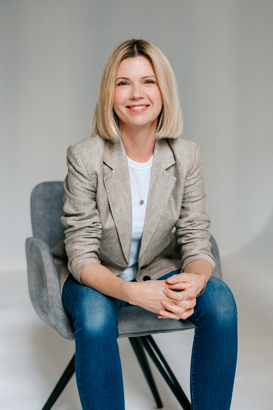 Eine Frau mit blondem, schulterlangem Haar sitzt auf einem grauen Stuhl und lächelt. Sie trägt einen beigen Blazer, ein weißes Oberteil, blaue Jeans und eine goldene Halskette und hat die Hände im Schoß gefaltet. Der Hintergrund ist einfarbig und hell.
