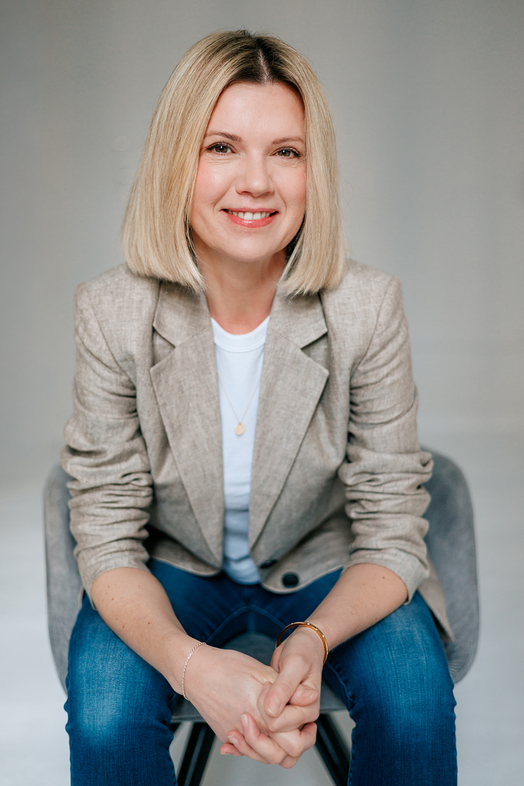 Eine Frau mit glattem blondem Haar, die einen beigen Blazer, ein weißes Hemd und eine blaue Jeans trägt, sitzt auf einem grauen Stuhl und lächelt in die Kamera, während sie die Hände vor sich verschränkt.