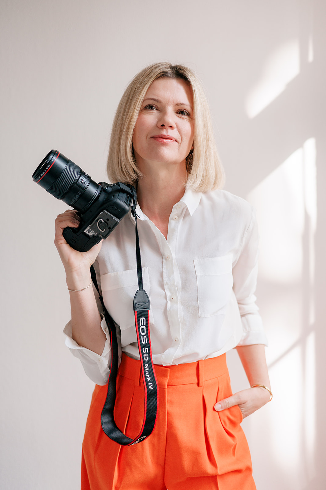 Eine Frau mit blondem Bobhaarschnitt, weißer Bluse und leuchtend orangefarbener Hose hält eine professionelle Kamera über die Schulter und lächelt selbstbewusst vor einem hellen Hintergrund.