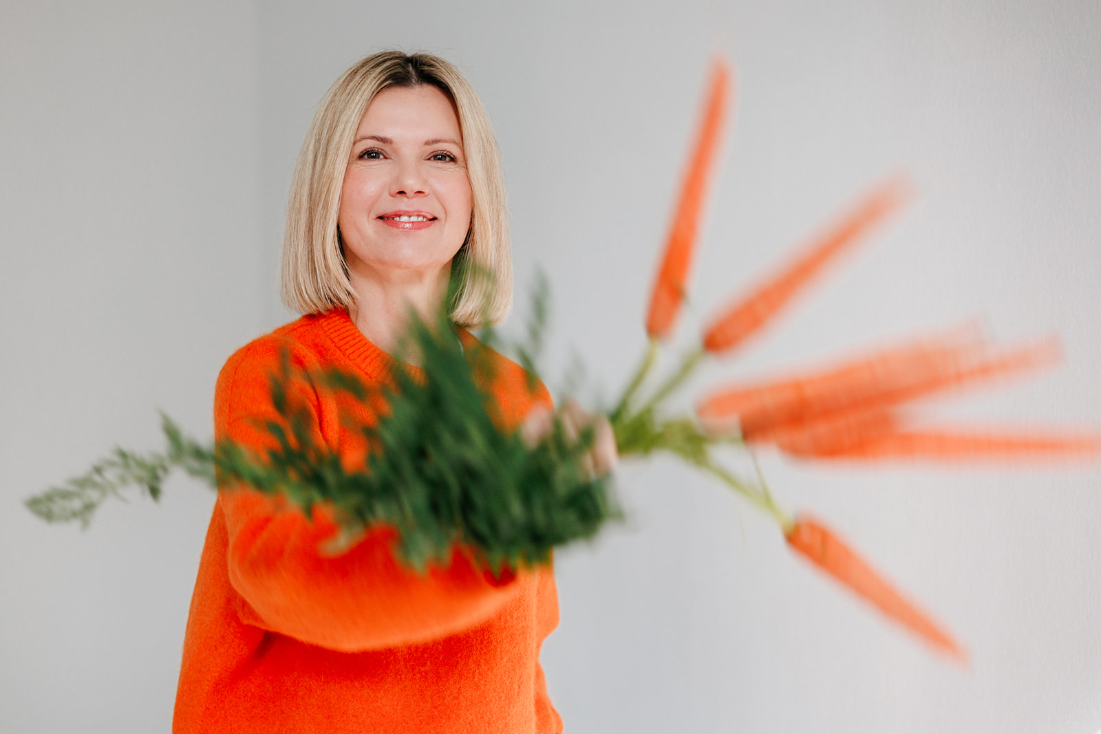 Eine lächelnde Frau in einem orangefarbenen Pullover hält einen Strauß frischer Karotten mit grünen Spitzen vor einem schlichten hellen Hintergrund.